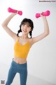 A woman in a yellow top and blue leggings holding two pink dumbbells.