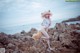 A woman in a white bikini and a straw hat on a rocky beach.