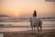 A woman in a white dress riding a white horse on the beach.